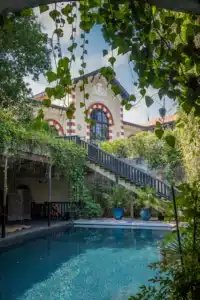 Piscine bleue scintillante, encadrée de végétation. En fond, bâtiment historique "COMPAGNIE GENERALE DES EAUX" aux arches rouges, avec escalier.