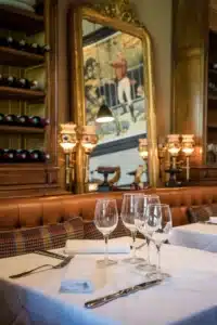 Table dressée dans un restaurant chic avec verres à vin, argenterie, banquette en cuir. Grand miroir doré reflète une peinture et des bouteilles.
