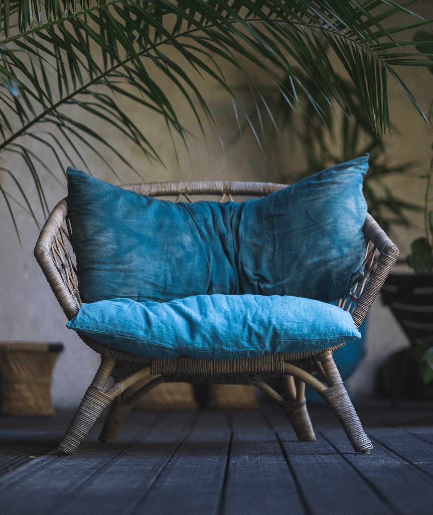 Fauteuil Rotin Bleu Coussin Ambiance Tropicale Fauteuil en rotin confortable avec coussins bleu sarcelle sous des feuilles de palmier luxuriantes, créant une ambiance tropicale et reposante.